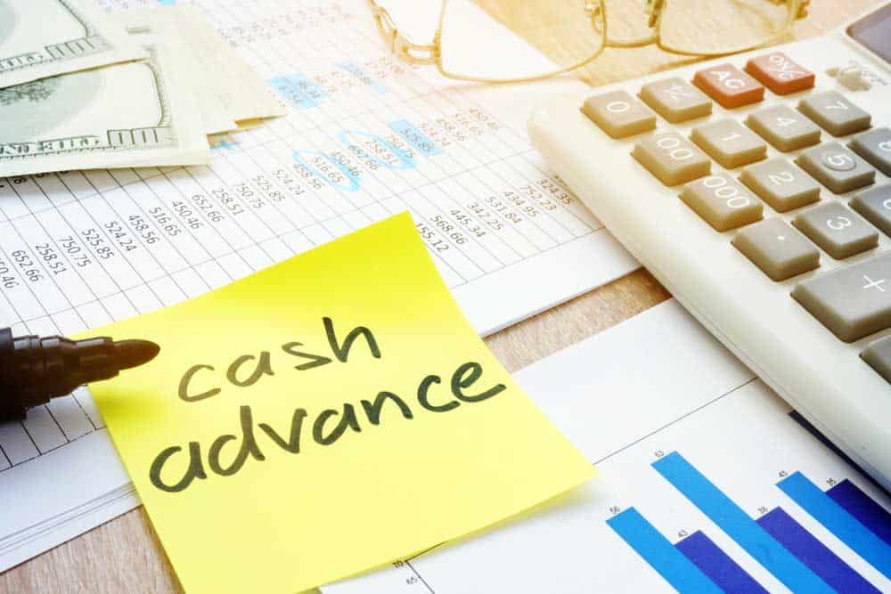 Post-it note with the words "Cash advance" on a table filled with papers, pens, glasses, calculator, and money