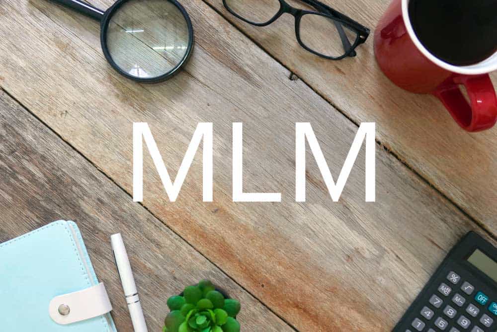 MLM written on top of a wooden tables containing a coffee mug, glasses, magnifying glass, calculater, notebook, and a pen.